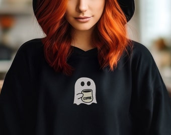 May include: A black hoodie featuring a white ghost holding a coffee mug with the word "Coffee" on it. The ghost is positioned on the front of the hoodie. The person wearing the hoodie has long red hair.