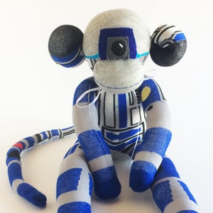 May include: A grey and blue sock monkey with a white ribbon tied around its neck. The monkey is sitting with its legs crossed and has a blue and grey striped pattern on its body.