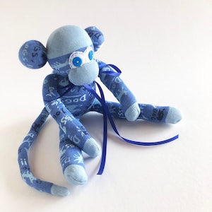 May include: A blue sock monkey with blue eyes and a blue ribbon tied around its neck. The monkey is sitting on a white surface.