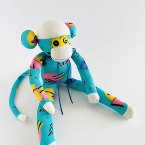 May include: A blue and pink sock monkey with a white face and yellow eyes. The monkey is sitting on a white surface.