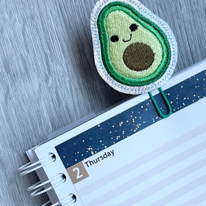 May include: A green avocado-shaped bookmark with a smiling face and a brown pit. The bookmark is attached to a green metal paper clip and is on top of a white and blue planner with the date "2 Thursday" written on it.