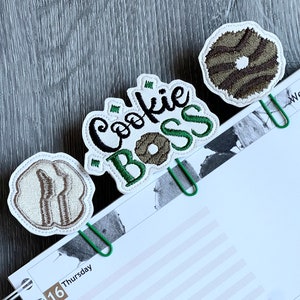 May include: Three green binder clips with embroidered patches. The patches are white with brown and green embroidery. One patch says "Cookie Boss" with a brown cookie in the middle. Another patch is a brown cookie with a white swirl. The third patch is a brown cookie with a white swirl.