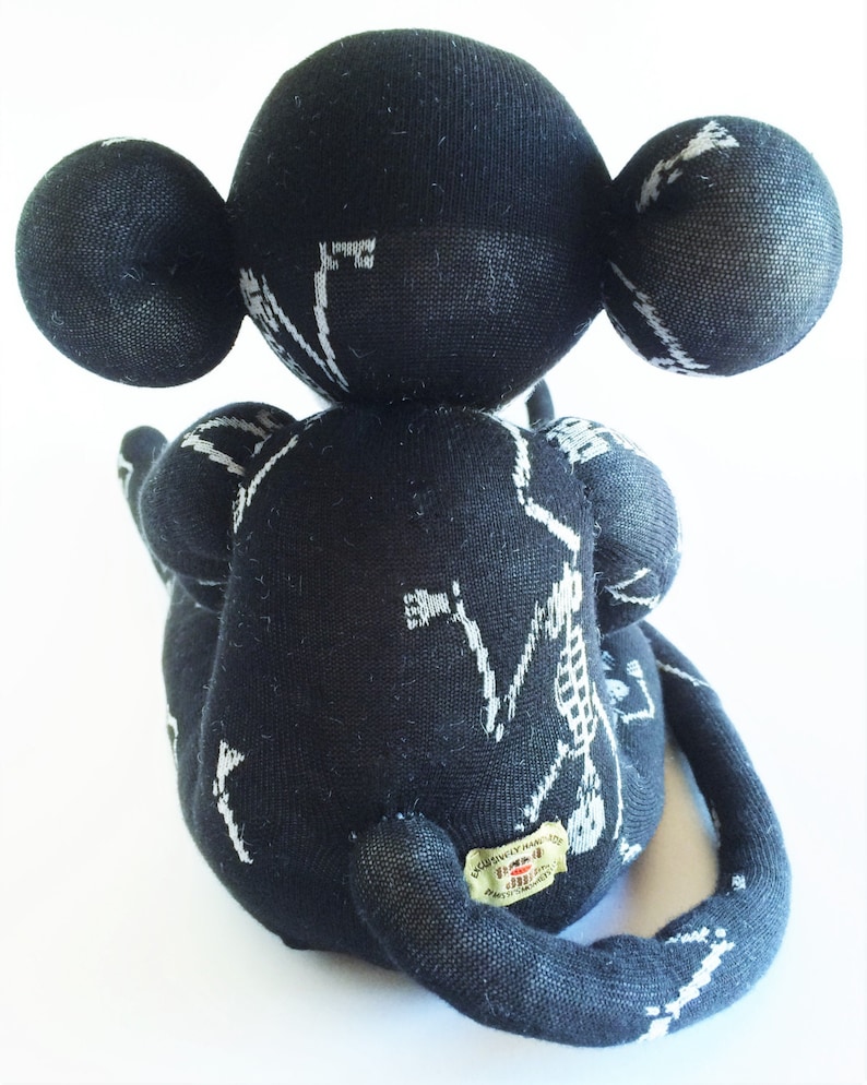 Puede incluir: Mono de calcet&iacute;n negro y blanco con un dise&ntilde;o de esqueleto. El mono est&aacute; sentado con la cola enroscada alrededor de su cuerpo. El mono tiene una etiqueta que dice "Exclusively Handmade by The Sock Monkey Shop".