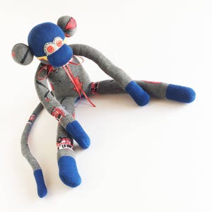 Puede incluir: Un mono de calcetín gris con pies azules y una cabeza azul. El mono tiene ojos rojos y una cinta roja alrededor del cuello. El mono está sentado sobre un fondo blanco.