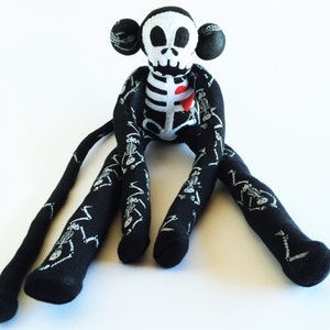 Puede incluir: Un mono de peluche esqueleto negro y blanco con un coraz&oacute;n rojo en el pecho. El mono tiene extremidades largas, como calcetines, con un patr&oacute;n de esqueleto.