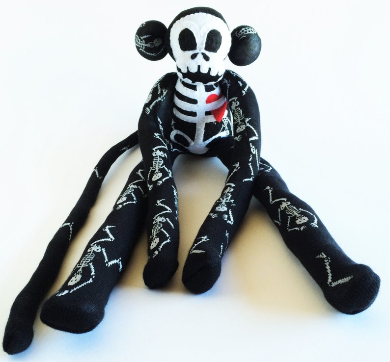 Puede incluir: Un mono de peluche esqueleto negro y blanco con un coraz&oacute;n rojo en el pecho. El mono tiene extremidades largas, como calcetines, con un patr&oacute;n de esqueleto.