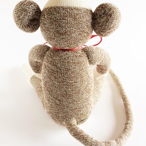 Puede incluir: Un mono de peluche de calcet&iacute;n marr&oacute;n y blanco con una pajarita roja. El mono est&aacute; sentado con la cola enroscada alrededor de su cuerpo.