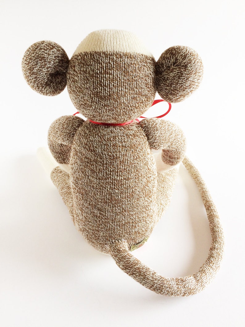 Puede incluir: Un mono de peluche de calcet&iacute;n marr&oacute;n y blanco con una pajarita roja. El mono est&aacute; sentado con la cola enroscada alrededor de su cuerpo.