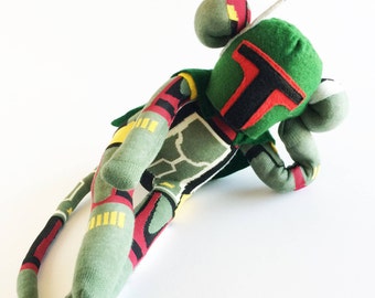 Puede incluir: Un juguete de peluche verde, rojo y amarillo con forma de personaje Boba Fett de Star Wars. El juguete tiene una cola larga y está tumbado de lado.