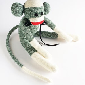 Könnte beinhalten: Ein grüner und weißer Socken-Affe mit einer schwarzen Schleife. Der Affe sitzt mit gekreuzten Beinen und seinem Schwanz um seinen Körper gewickelt.