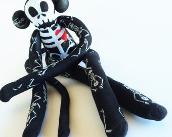 May include: A black and white skeleton monkey plush toy with a red heart on its chest. The monkey is sitting with its legs crossed and its arms folded.