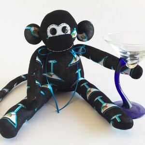 May include: A handmade sock monkey with a black body and head, holding a martini glass. The monkey's body is patterned with martini glasses in blue and white. The martini glass has a blue stem and a clear bowl.