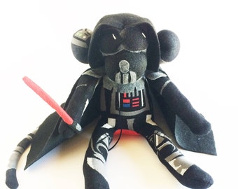 Puede incluir: Un mono de peluche negro y gris vestido como Darth Vader de Star Wars. El mono lleva una capa negra y un casco negro con un sable de luz rojo en la mano.