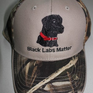 May include: A gray and camouflage baseball cap with an embroidered black Labrador Retriever and the text "Black Labs Matter".