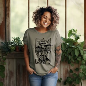 May include: Olive green t-shirt with a black illustration of a woman in a witch hat holding a cauldron, and the words "Kitchen Witch". The tee shirt is made of a soft material and is suitable for casual wear.
