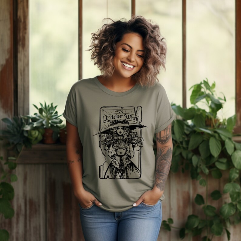 May include: Olive green t-shirt with a black illustration of a woman in a witch hat holding a cauldron, and the words "Kitchen Witch". The tee shirt is made of a soft material and is suitable for casual wear.