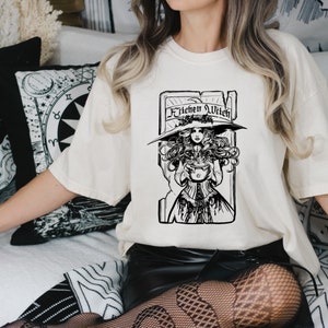 May include: A cream-colored t-shirt featuring a black and white illustration of a witch holding a cauldron. The text "Kitchen Witch" is above the witch. The witch is wearing a large hat and a long dress. The shirt is a loose fit.