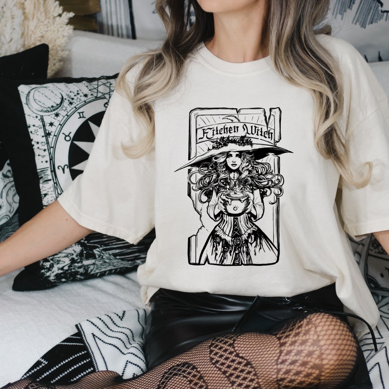 May include: A cream-coloured t-shirt with a black and white illustration of a witch holding a cauldron. The text "Kitchen Witch" is above the witch. The witch is wearing a large hat and a long dress. The shirt has a loose fit.