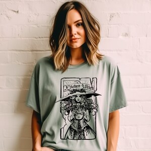 May include: Sage green t-shirt with a black and white illustration of a witch holding a cauldron. The witch is wearing a large hat and is framed by a rectangular border with the words "Kitchen Witch" across the top.