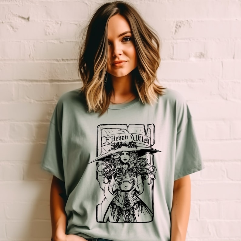 May include: Sage green t-shirt with a black and white illustration of a witch holding a cauldron. The witch is wearing a large hat and is framed by a rectangular border with the words "Kitchen Witch" across the top.