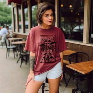 May include: A maroon t-shirt with a black graphic of a witch and the words "Kitchen Witch". The witch is wearing a large hat and holding a cauldron. The t-shirt has a relaxed fit.