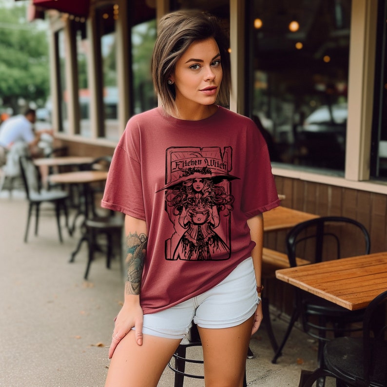 May include: A maroon t-shirt with a black graphic of a witch and the words "Kitchen Witch". The witch is wearing a large hat and holding a cauldron. The t-shirt has a relaxed fit.