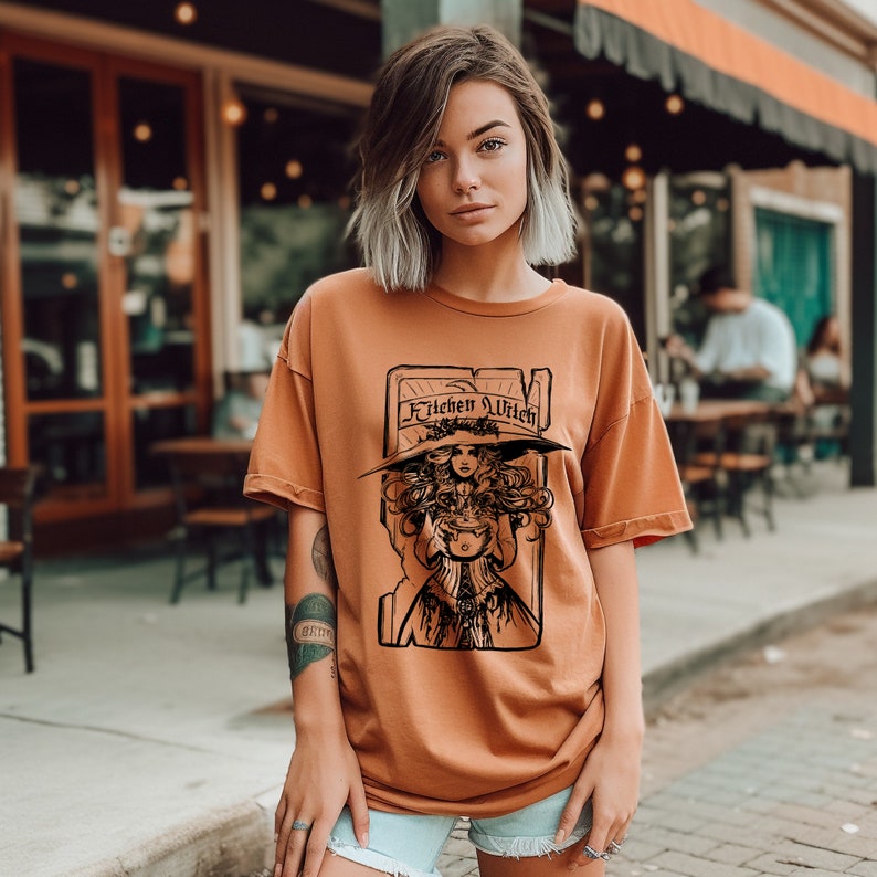 May include: Orange-brown t-shirt featuring a black graphic of a woman in a witch hat holding a cauldron, with the text "Kitchen Witch" above. The shirt has a relaxed fit.