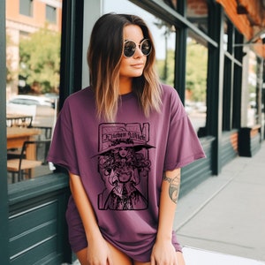 May include: A plum-coloured t-shirt with a black graphic of a witch and the text "Kitchen Witch". The witch is wearing a large hat and holding a cauldron. The shirt has a relaxed fit and short sleeves.