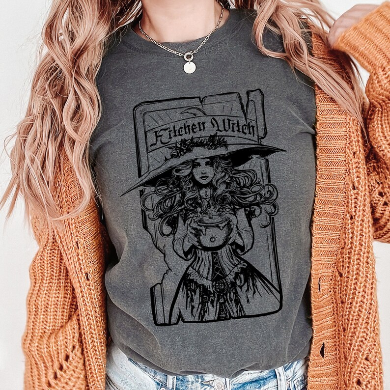 May include: Dark grey t-shirt with a black graphic of a woman in a witch hat holding a cauldron, with the words "Kitchen Witch" above. The woman is wearing a long dress. The shirt is worn with a rust-coloured cardigan.