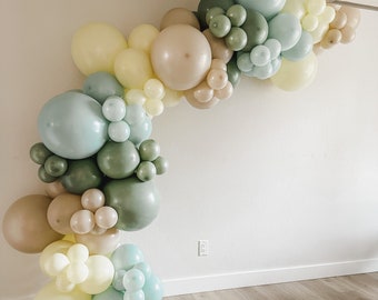 May include: A balloon arch made of light blue, beige, and green balloons. The arch is arranged in a cascading pattern and is attached to a white wall.