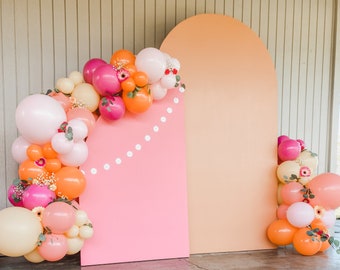 May include: A party backdrop featuring a pink rectangular panel and a peach-colored arch. A balloon garland in shades of pink, peach, and orange, adorned with flowers, frames the pink panel. The backdrop is set on a concrete surface.