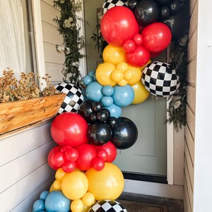 May include: A colorful balloon garland in red, yellow, black, blue, and checkered patterns adorns a doorway. The arrangement includes various sizes of balloons, creating a festive and celebratory atmosphere. Greenery adds a natural touch to the decoration.