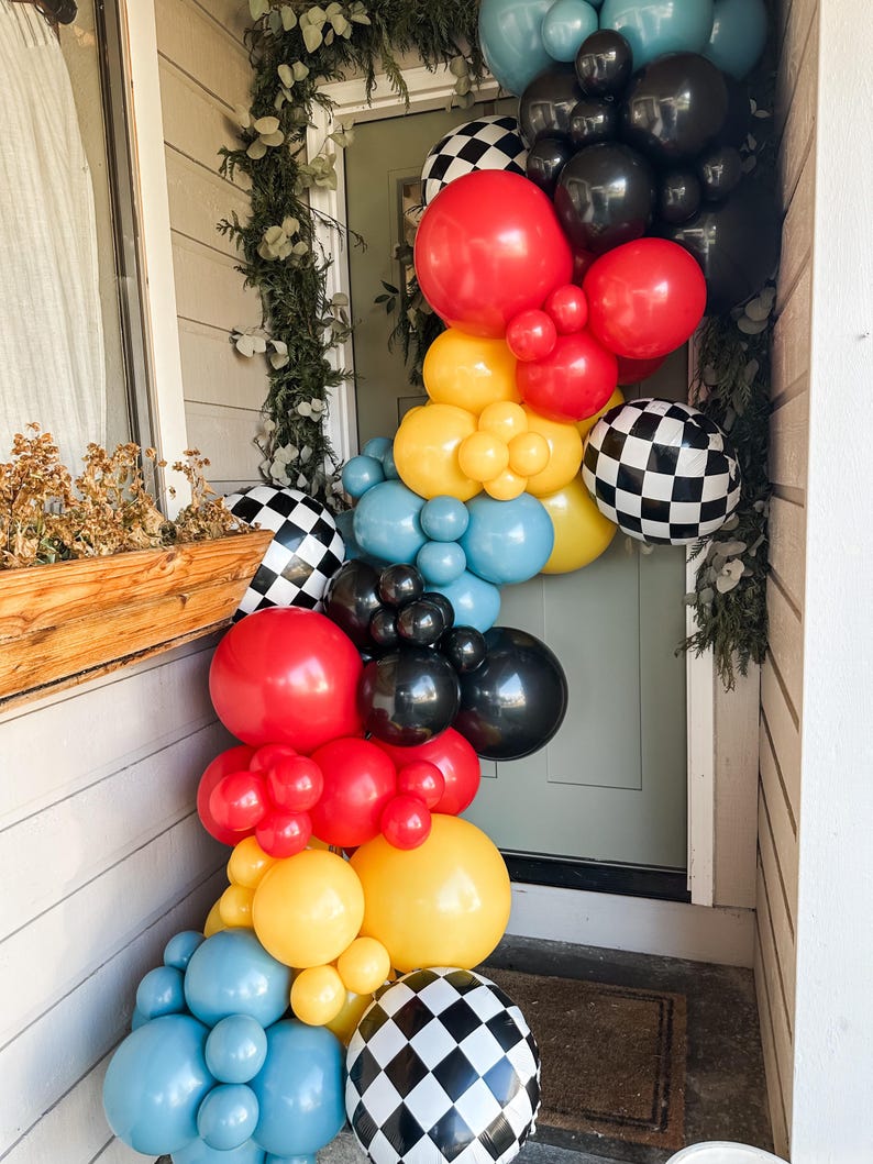 May include: A colorful balloon garland in red, yellow, black, blue, and checkered patterns adorns a doorway. The arrangement includes various sizes of balloons, creating a festive and celebratory atmosphere. Greenery adds a natural touch to the decoration.