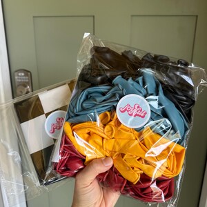 May include: A person holding two clear plastic bags of balloons. One bag contains a black and white checkered pattern. The other bag contains a mix of colorful balloons: black, teal, yellow, and red. Each bag has a sticker that says "joyfull".