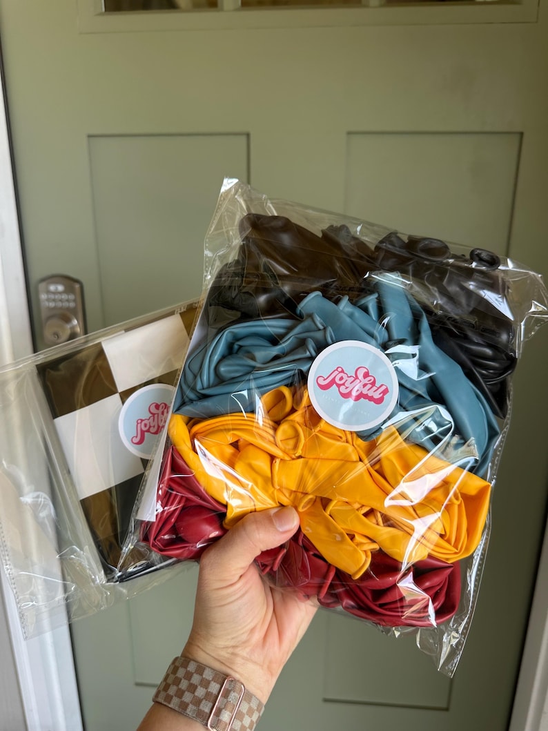 May include: A person holding two clear plastic bags of balloons. One bag contains a black and white checkered pattern. The other bag contains a mix of colorful balloons: black, teal, yellow, and red. Each bag has a sticker that says "joyfull".
