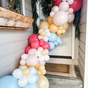 Può includere: Una ghirlanda di palloncini realizzata con palloncini rosa pastello, gialli, blu e rossi. La ghirlanda è decorata con palloncini bianchi a forma di fiori. La ghirlanda è appesa a un muro bianco accanto a una porta.