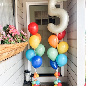 May include: Two balloon bouquets featuring a large cream-colored number five foil balloon and various latex balloons in red, orange, yellow, light green, blue, and dark blue. The balloons are arranged in clusters with smaller balloons at the base.