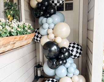 May include: A balloon garland featuring a mix of matte black, light blue, and beige balloons, interspersed with black and white checkered pattern balloons. The arrangement is vertically oriented, cascading down the side of a white door frame.