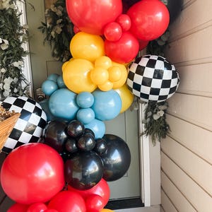 May include: A vibrant balloon garland in red, yellow, blue, black, and checkered patterns. The balloons are arranged in clusters, creating a festive and eye-catching display. The garland is likely for a party or celebration.
