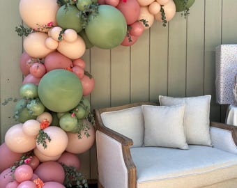 May include: A balloon arch in shades of pink, green, and peach, adorned with small flowers and greenery. The balloons are arranged in a cascading design, creating a festive and decorative display. A light-colored armchair with two pillows is in the background.