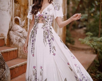 May include: A woman wearing a white maxi dress with a floral print. The dress has a V-neckline and short sleeves. The woman is standing on a set of stairs with a stone wall in the background.