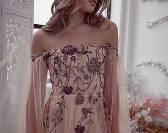Pode incluir: Vestido cor de rosa pêssego com bordado floral. O vestido apresenta um corpete justo e mangas de tule esvoaçantes. O design floral incorpora tons de roxo e verde.