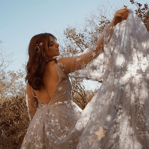 May include: A woman wearing a light blue tulle dress with gold star embellishments. The dress has a low back and long sleeves. The woman is holding the fabric of the dress in her hand, creating a flowing effect.