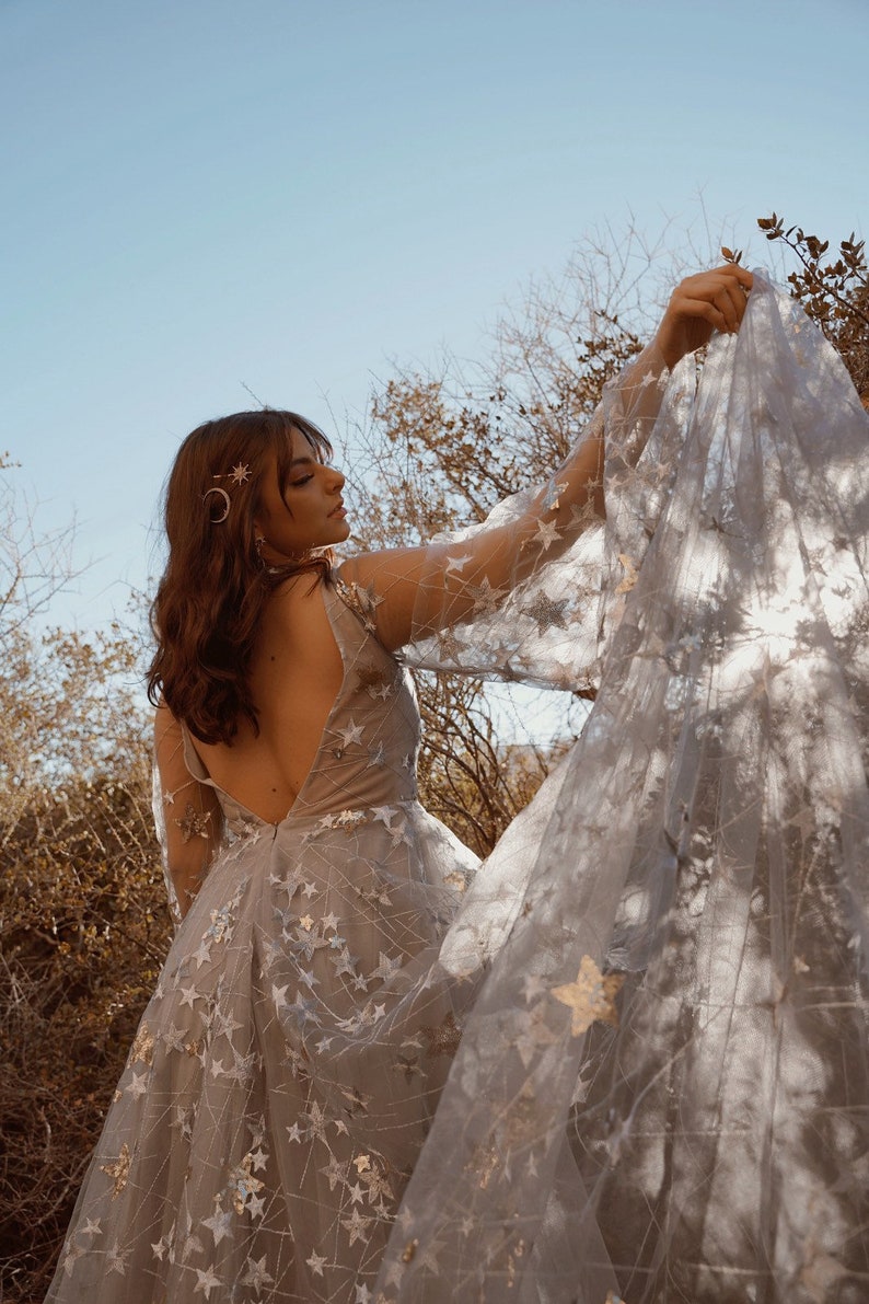 May include: A woman wearing a light blue tulle dress with gold star embellishments. The dress has a low back and long sleeves. The woman is holding the fabric of the dress in her hand, creating a flowing effect.