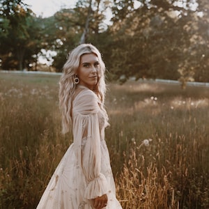 May include: A woman in a long, flowing, off-white gown with gold star and moon embroidery stands in a field of tall grass. The gown has long, sheer sleeves with ruched detailing at the cuffs. The woman has long, blonde hair and is looking over her shoulder.