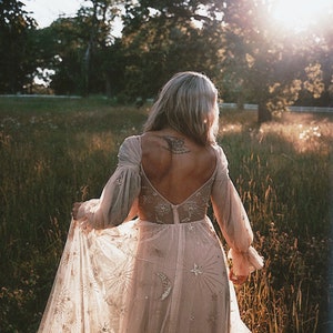 May include: A woman wearing a long, flowing, light pink dress with a star and moon pattern. The dress has a low back and long sleeves. The woman is standing in a field of tall grass with trees in the background.