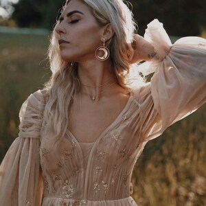 May include: A woman wearing a beige tulle dress with gold star embellishments. She is wearing gold earrings and a gold necklace with a star pendant. The dress has long sleeves with a gathered detail at the shoulders.