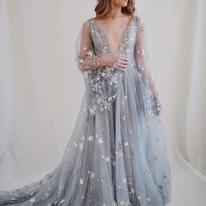 May include: A long, flowing, light blue tulle gown with a plunging neckline and long sleeves. The gown is covered in silver and gold stars.