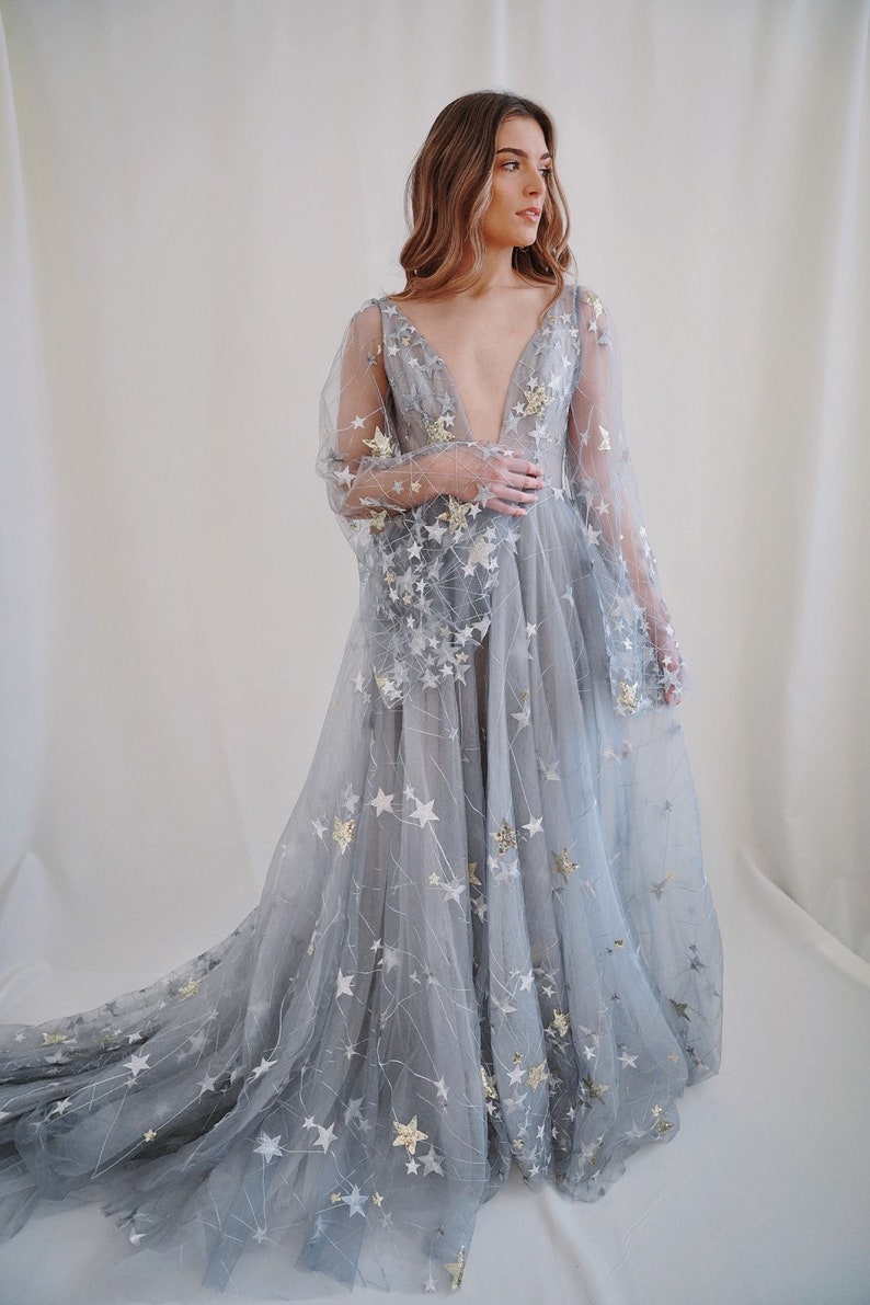 May include: A long, flowing, light blue tulle gown with a plunging neckline and long sleeves. The gown is covered in silver and gold stars.