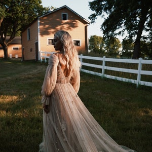 May include: A woman in a long, flowing, beige tulle gown with a delicate floral pattern and a low back. The gown has long sleeves with puffed shoulders. She is standing in a grassy field with a white fence and a barn in the background.
